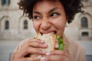 Close up shot of young curly woman feels hungry eats sandwich with appetite poses at street focused somewhere stands outdoors against blurred background prefers junk food. Nutrition concept